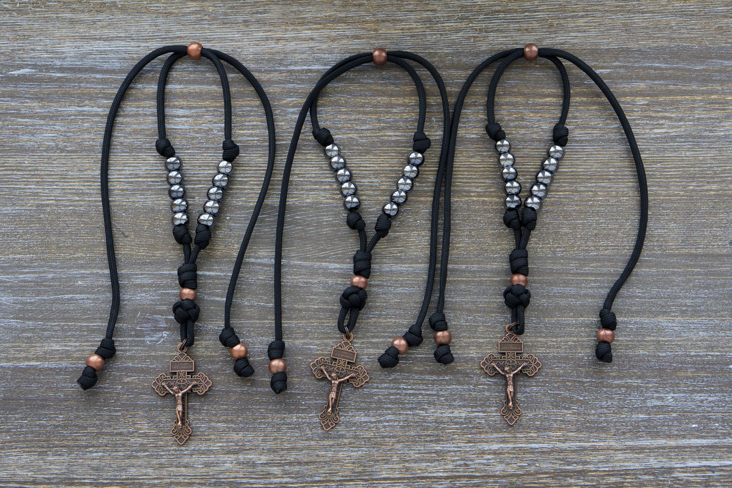 Black paracord and antique copper rosary hanging from a car rearview mirror. Features gunmetal Hail Mary beads, a 2-inch antique copper Pardon Crucifix.