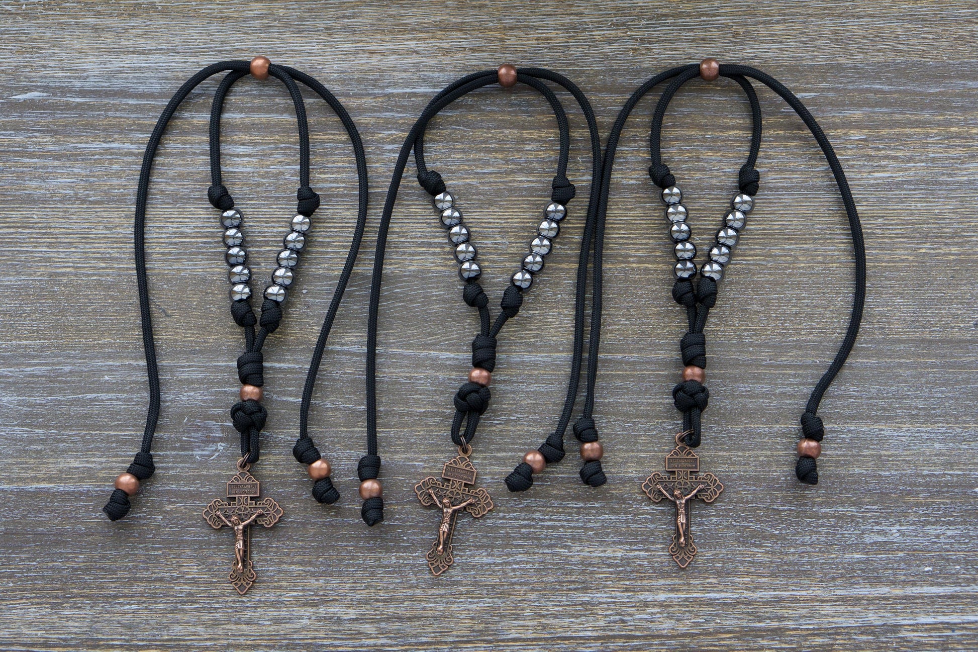 Black paracord and antique copper rosary hanging from a car rearview mirror. Features gunmetal Hail Mary beads, a 2-inch antique copper Pardon Crucifix.