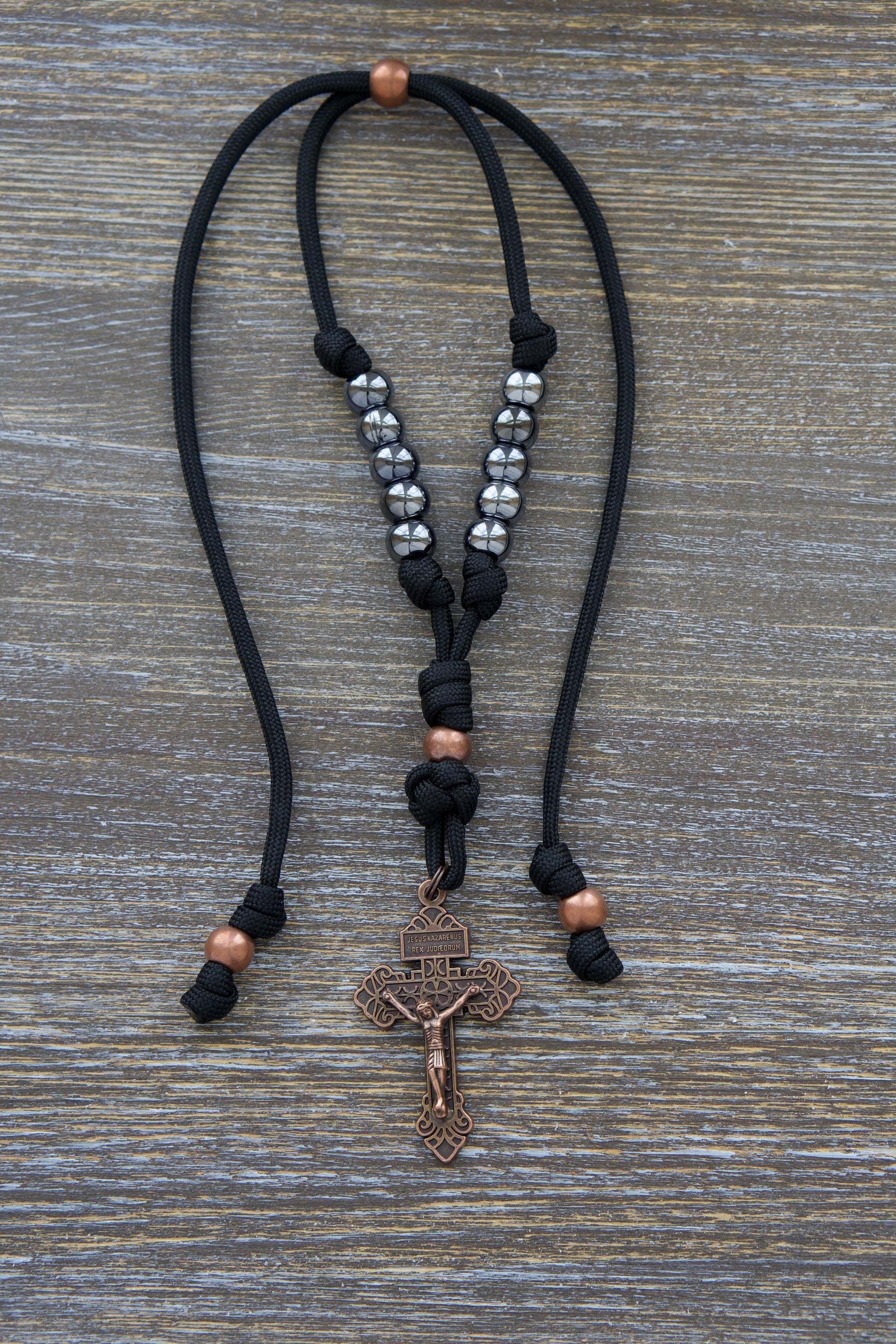 Black paracord and antique copper rearview mirror rosary, featuring gunmetal Hail Mary beads and a 2-inch antique copper Pardon crucifix. Designed for durability and faith-inspired strength while driving.
