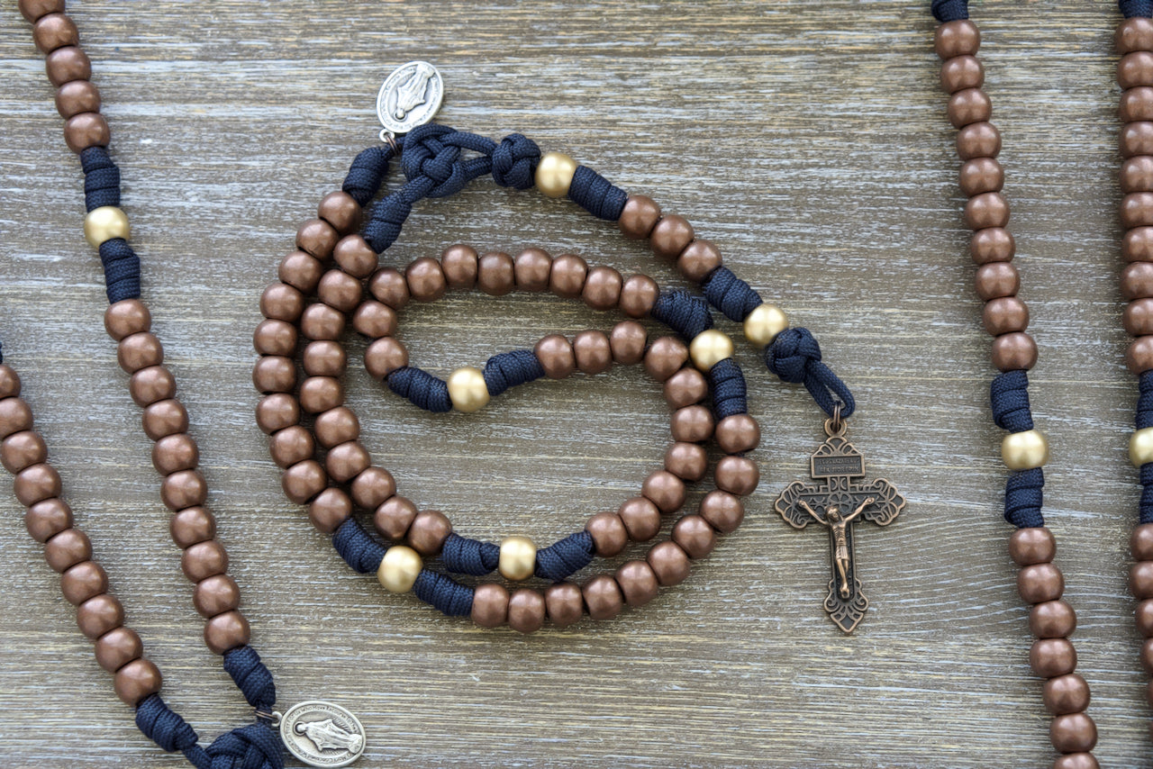 Exquisite 5 decade paracord rosary with navy blue rope, antique copper Hail Mary beads, and matte gold Our Father beads. Includes a 2-inch copper Pardon Crucifix, Miraculous Medal, and is handmade by a small Catholic family.