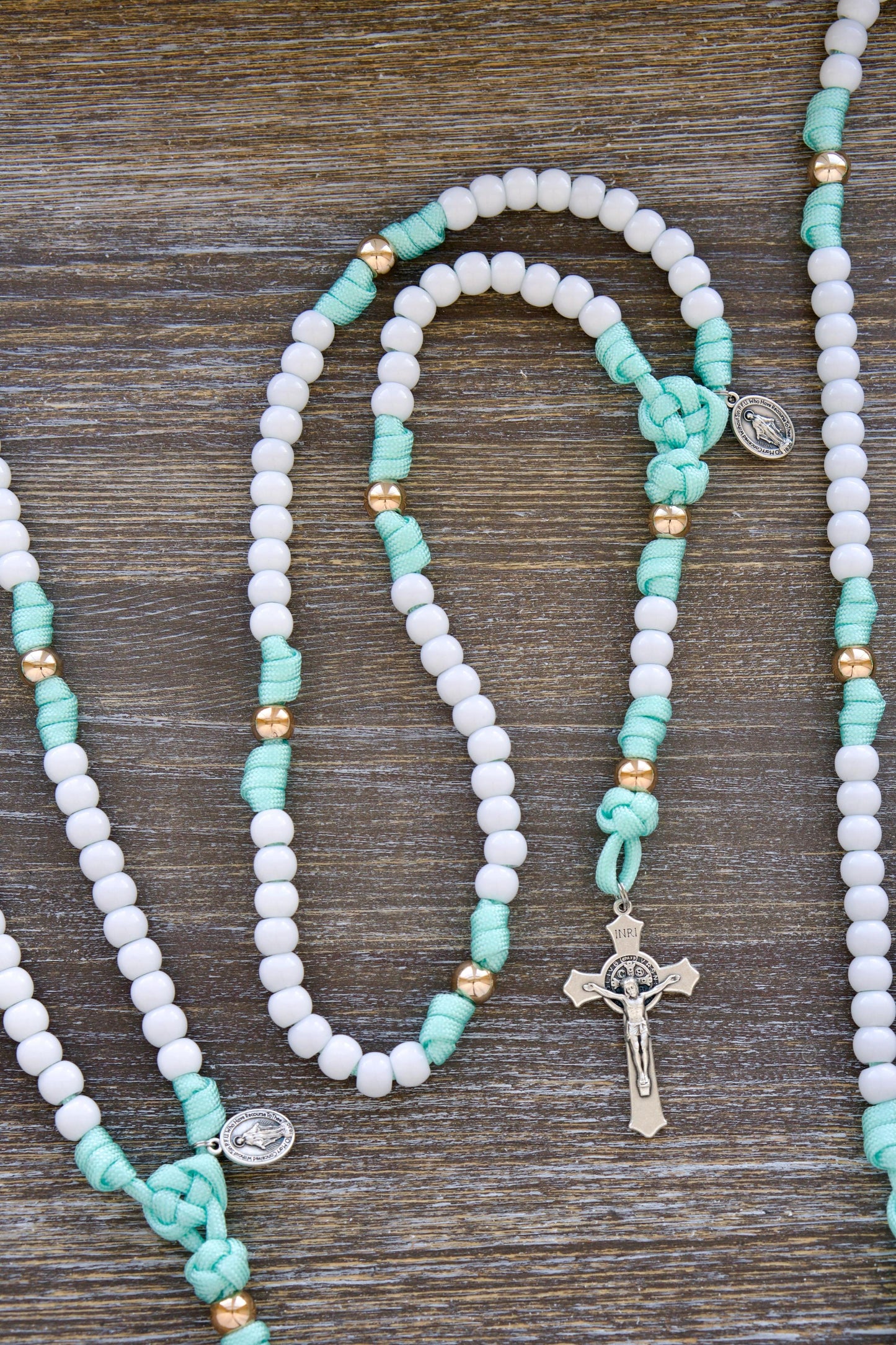 A mint green and white paracord rosary designed for children, featuring smaller 10mm acrylic beads, a rose gold Our Father bead accent, and a miniature Miraculous Medal and St. Benedict crucifix.