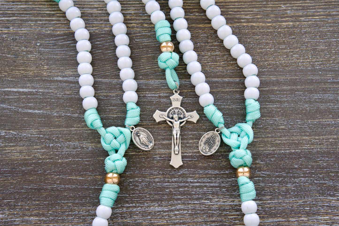 Close-up image of a mint green and white paracord rosary designed for children. Features durable 10mm acrylic beads, a rose gold Our Father bead accent, smaller Miraculous Medal, and a 2-inch St. Benedict crucifix.