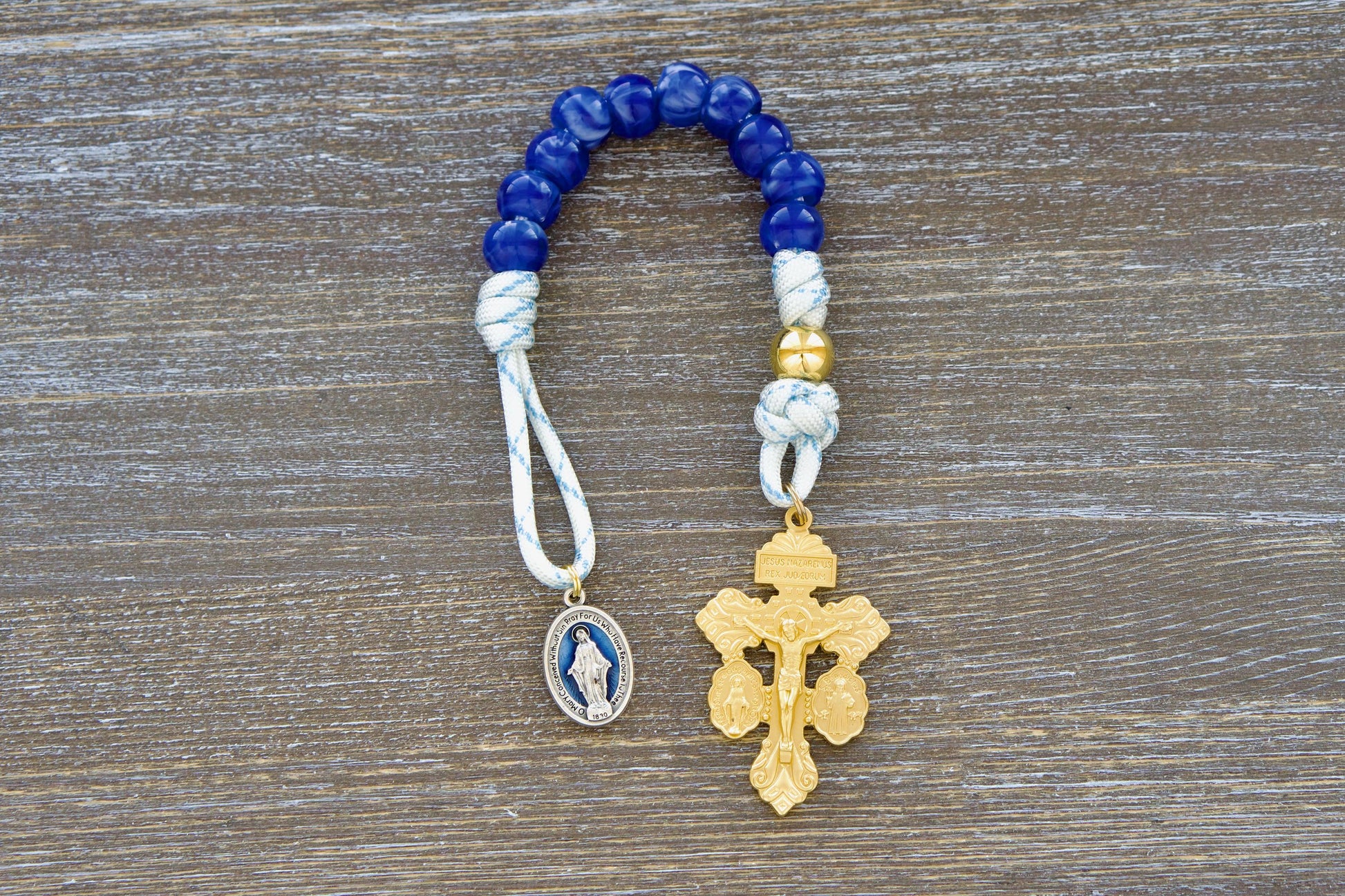 A single decade white paracord rosary, the Divine Light, features blue accents, a gold Our Father bead, custom blue 'miraculous blue' acrylic Hail Mary beads, a blue enamel Miraculous medal, and a gold 'triple threat' pardon crucifix. Handmade with durable 550 paracord.