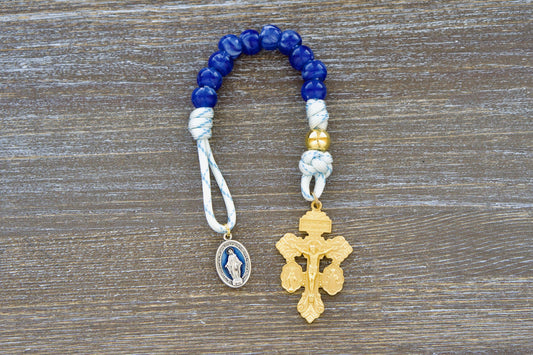 A single decade white paracord rosary, the Divine Light, features blue accents, a gold Our Father bead, custom blue 'miraculous blue' acrylic Hail Mary beads, a blue enamel Miraculous medal, and a gold 'triple threat' pardon crucifix. Handmade with durable 550 paracord.