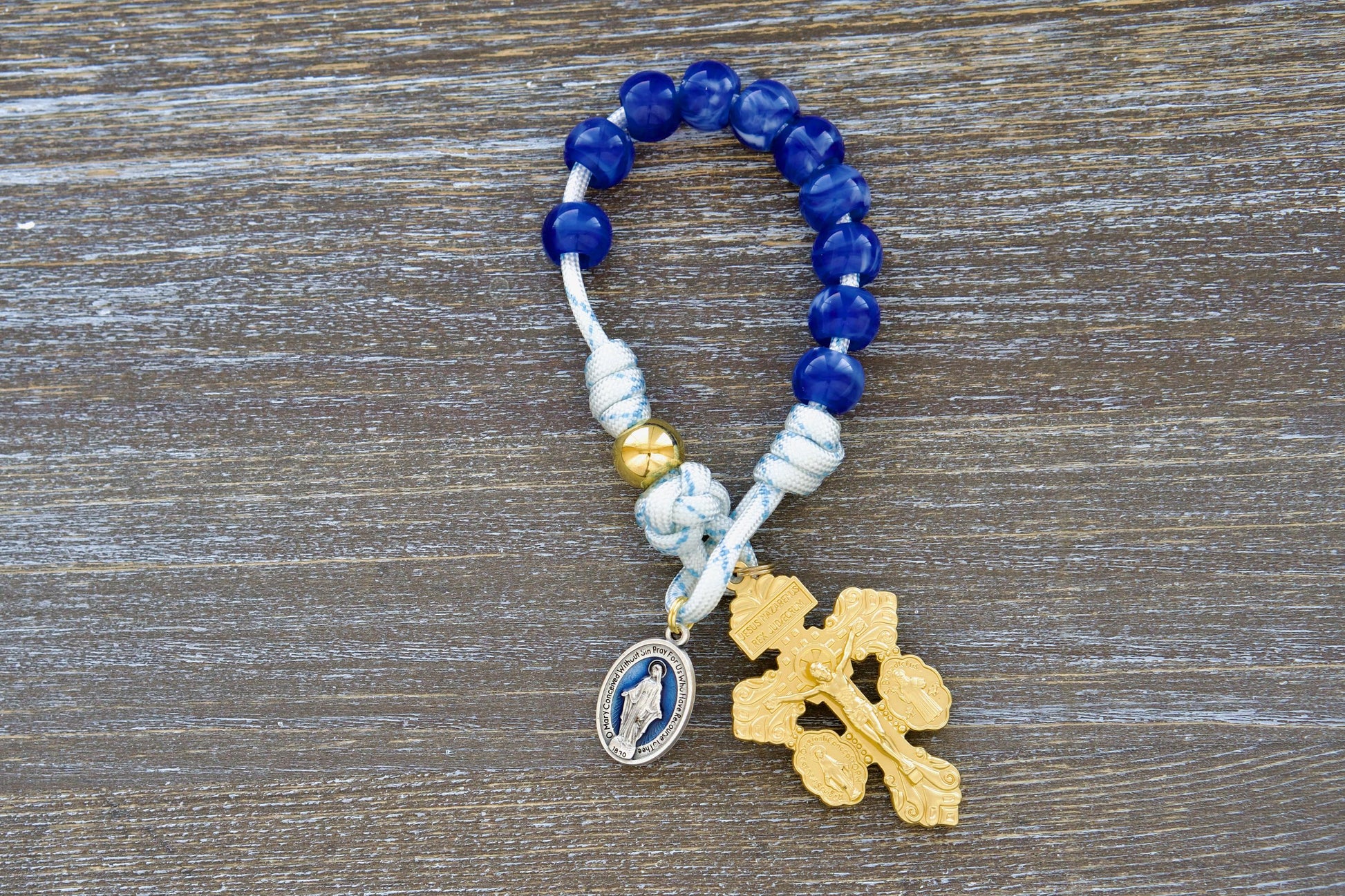 A single decade, white paracord rosary with blue accents, featuring a gold Our Father bead, custom blue acrylic Hail Mary beads, a blue enamel Miraculous medal, and a golden 'triple threat' pardon crucifix. Designed for portability, this 'Divine Light' pocket rosary from Sanctus Servo embodies strength, durability, and devotion.
