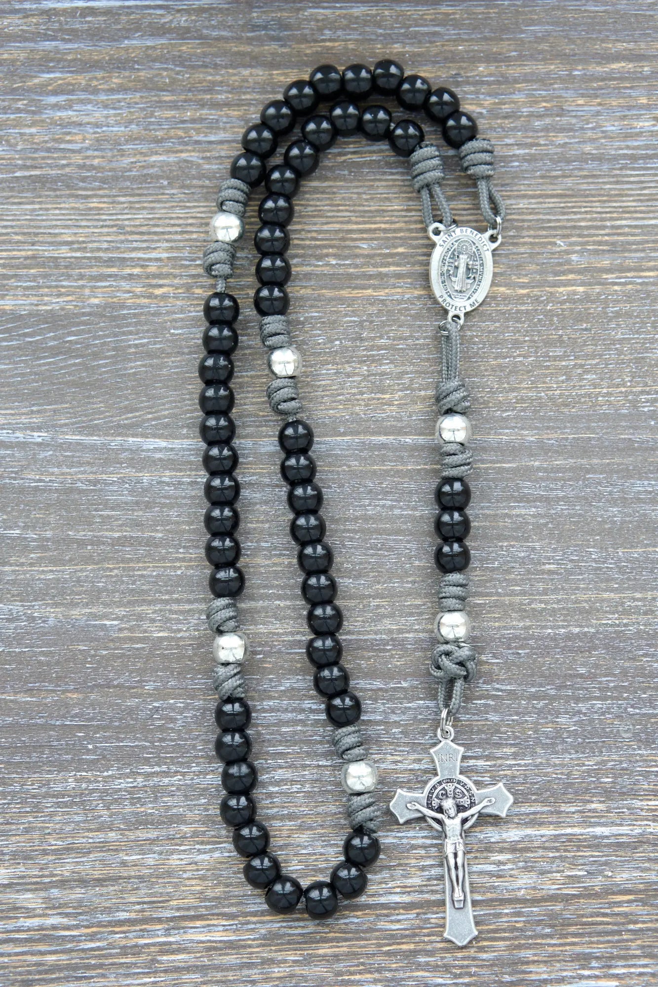 Silver Our Father beads accented by black metal Hail Mary beads on an iron gray paracord rosary with a St. Benedict crucifix.