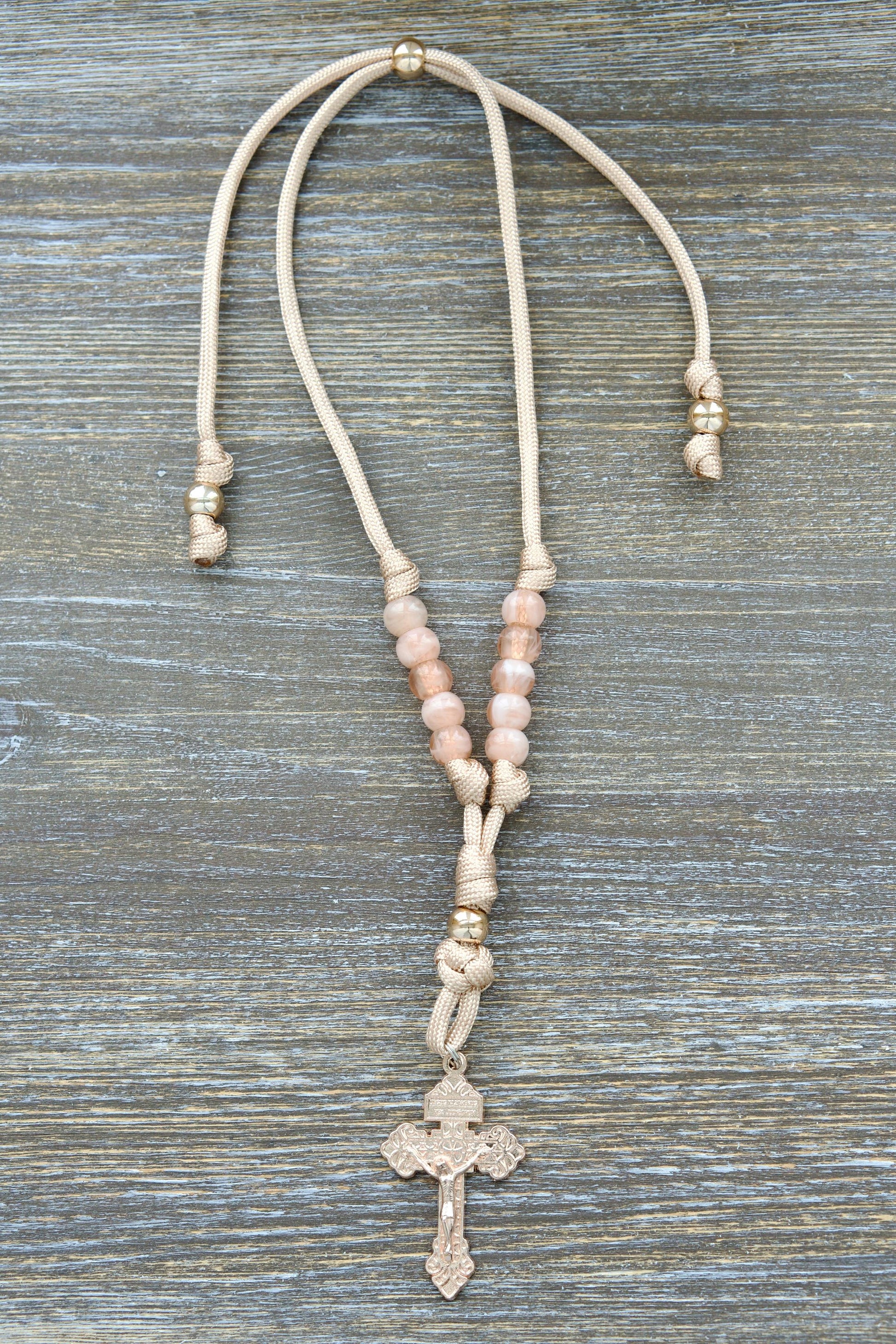 Tan and rose gold paracord rearview mirror rosary with milky pink Hail Mary beads, a striking rose gold Our Father bead, and a unique shop-exclusive rose gold Pardon crucifix centerpiece. Handmade by Sanctus Servo.