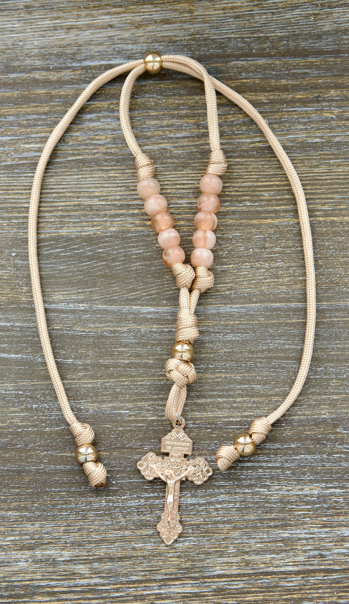 Gate of Heaven Rearview Mirror Rosary: A handcrafted tan and rose gold paracord rosary featuring milky pink Hail Mary beads, a striking rose gold Our Father bead, and an exclusive rose gold Pardon Crucifix.