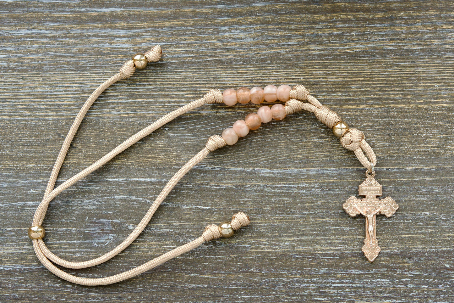 Tan and rose gold paracord rosary hanging from a rearview mirror. Features rose gold Our Father bead, milky pink Hail Mary beads, and a unique rose gold Pardon crucifix centerpiece.
