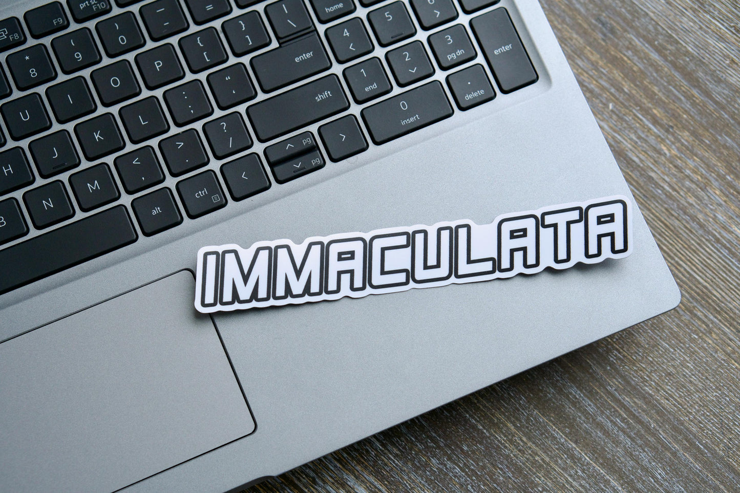Immaculata Vinyl Sticker - A white semi-glossy sticker (7.17" x 1.14") featuring the Militia of the Immaculata emblem, honoring St. Maximilian Kolbe's mission against heresy and modernism. Matte laminated for durability and UV resistance, suitable for indoor/outdoor use on various surfaces.