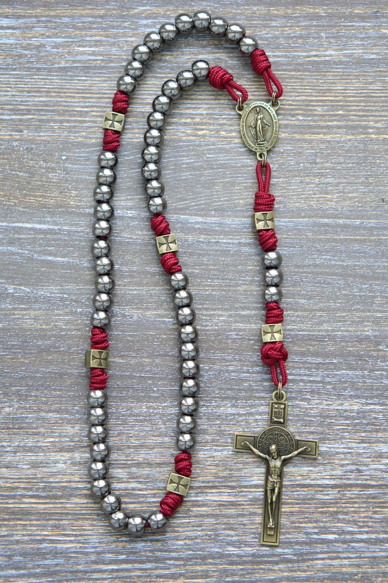 A handcrafted paracord rosary featuring gunmetal Hail Mary beads, an olive Miraculous Medal centerpiece, and a matching St. Benedict crucifix, crimson red rope.