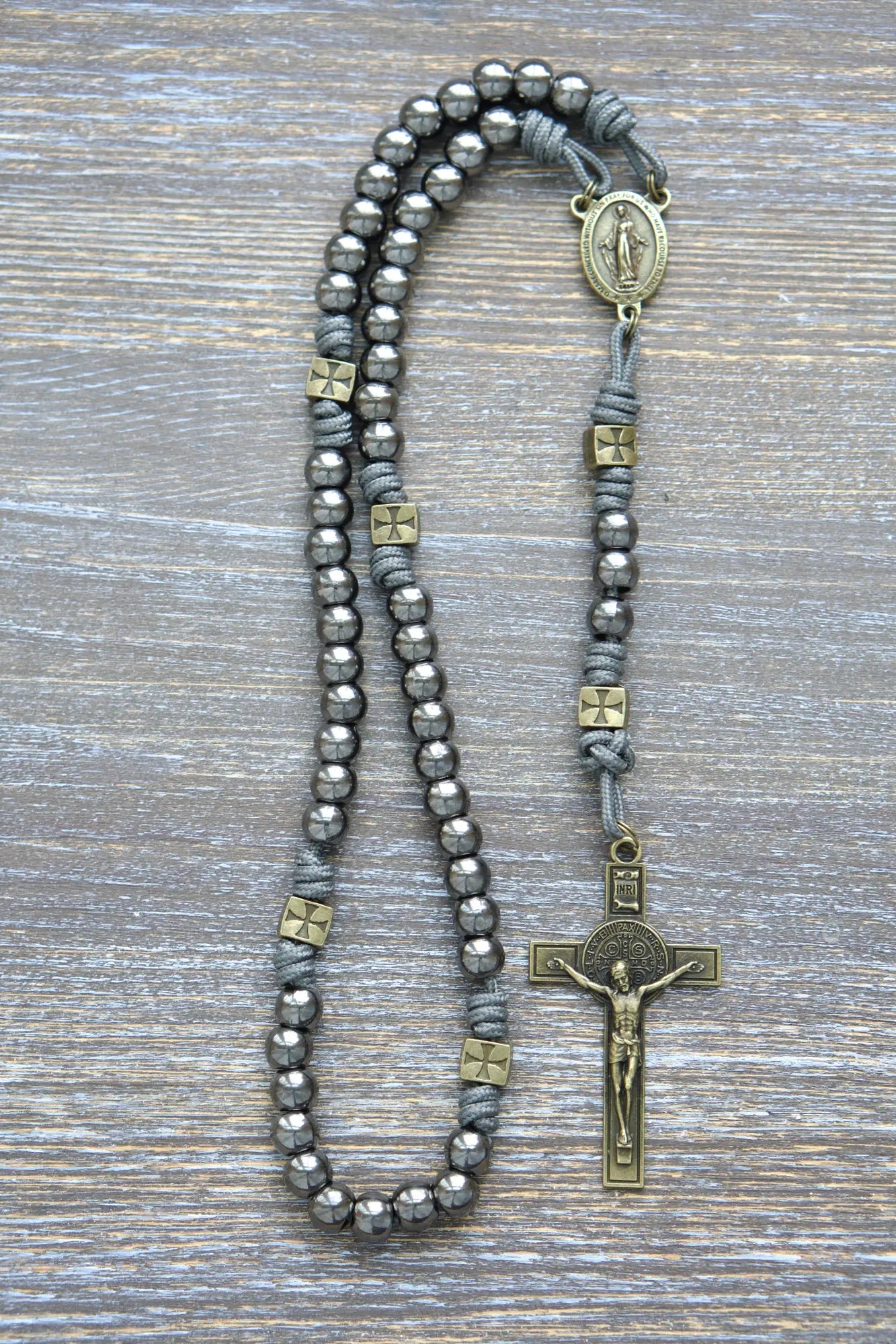 A handcrafted paracord rosary featuring gunmetal Hail Mary beads, an olive Miraculous Medal centerpiece, and a matching St. Benedict crucifix, iron gray rope.