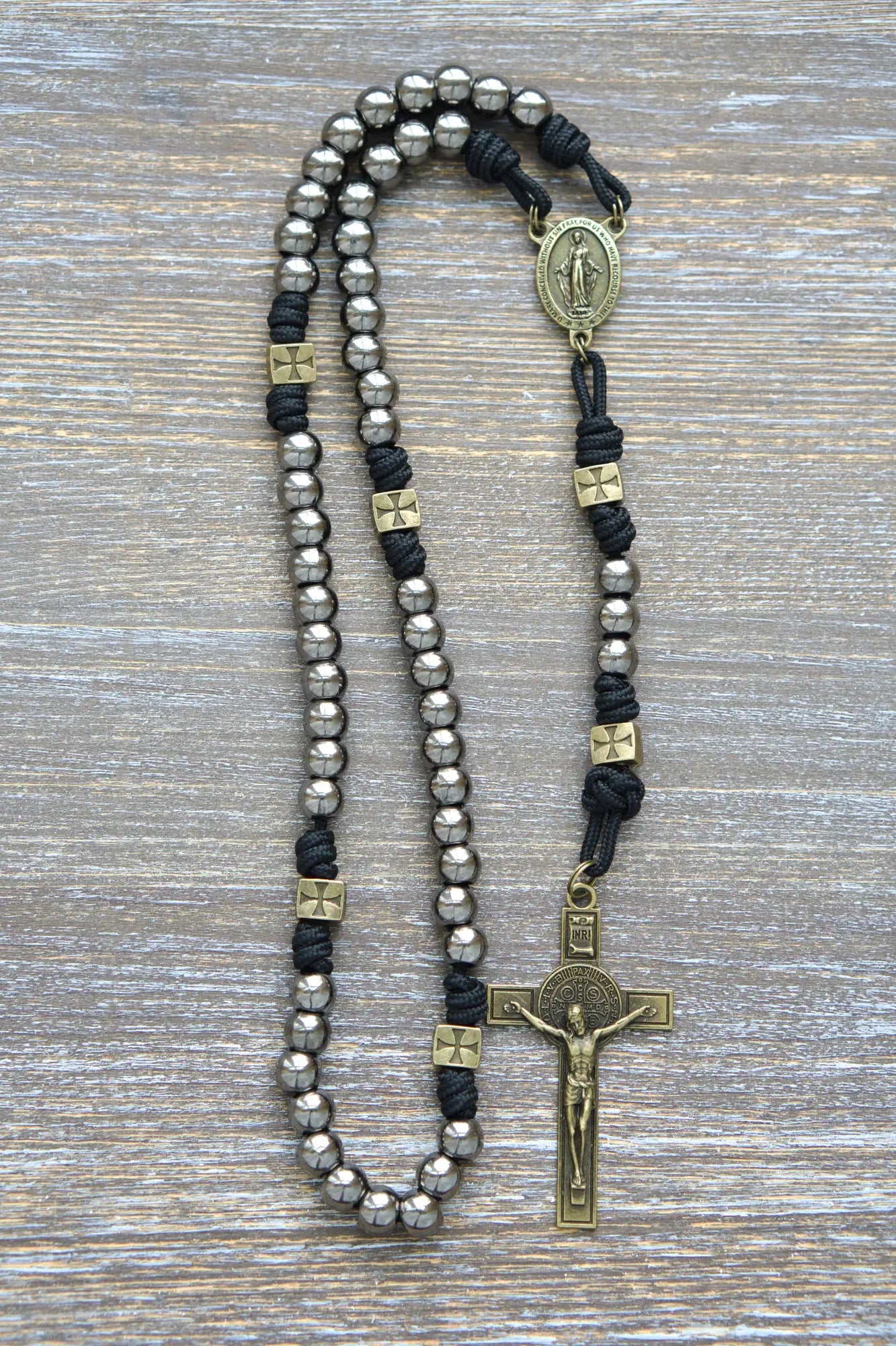 A handcrafted paracord rosary featuring gunmetal Hail Mary beads, an olive Miraculous Medal centerpiece, and a matching St. Benedict crucifix, midnight black rope.
