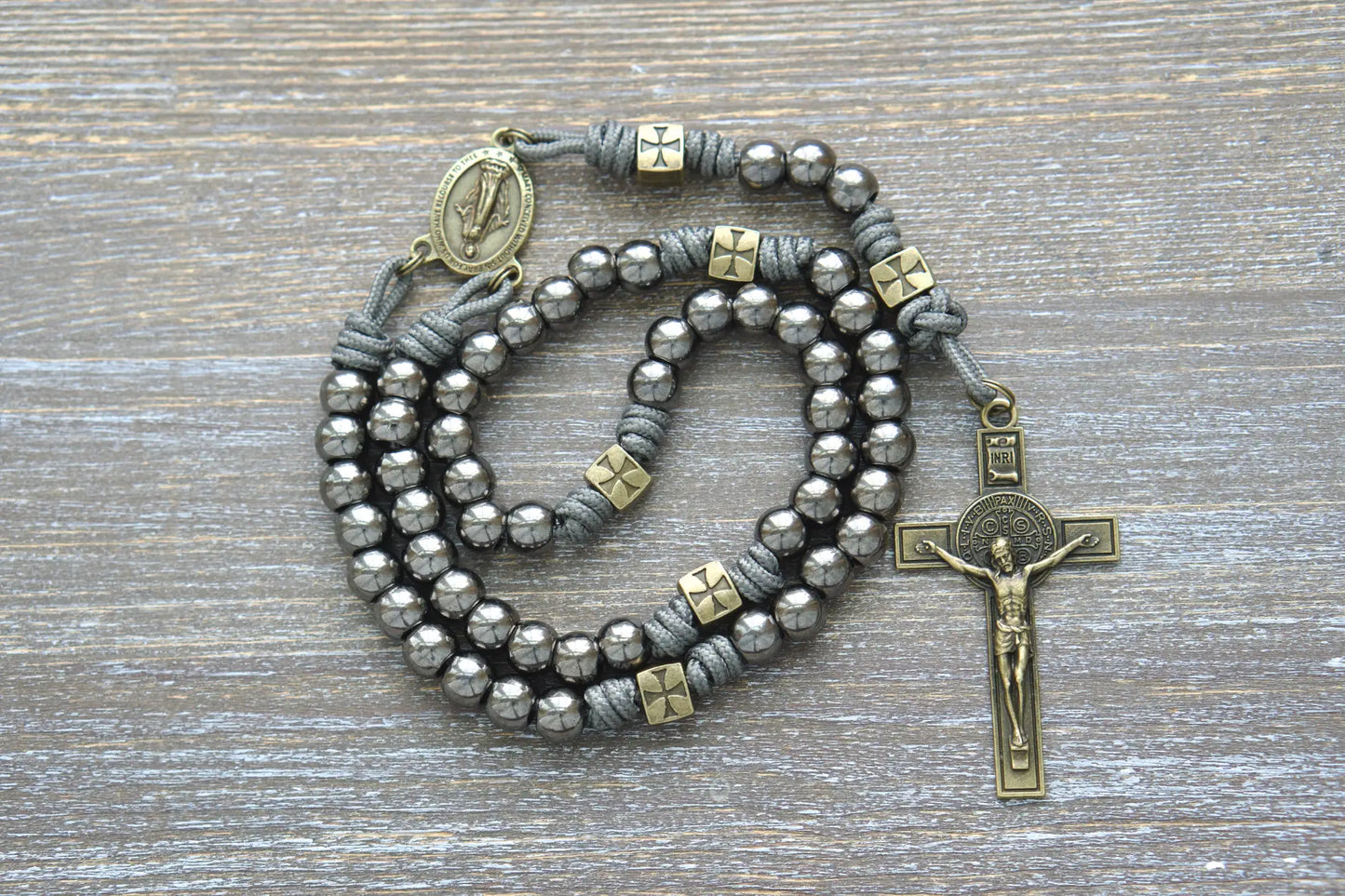 A rugged metal paracord rosary featuring olive Our Father beads, gunmetal Hail Mary beads, a Miraculous Medal centerpiece, and a St. Benedict crucifix.