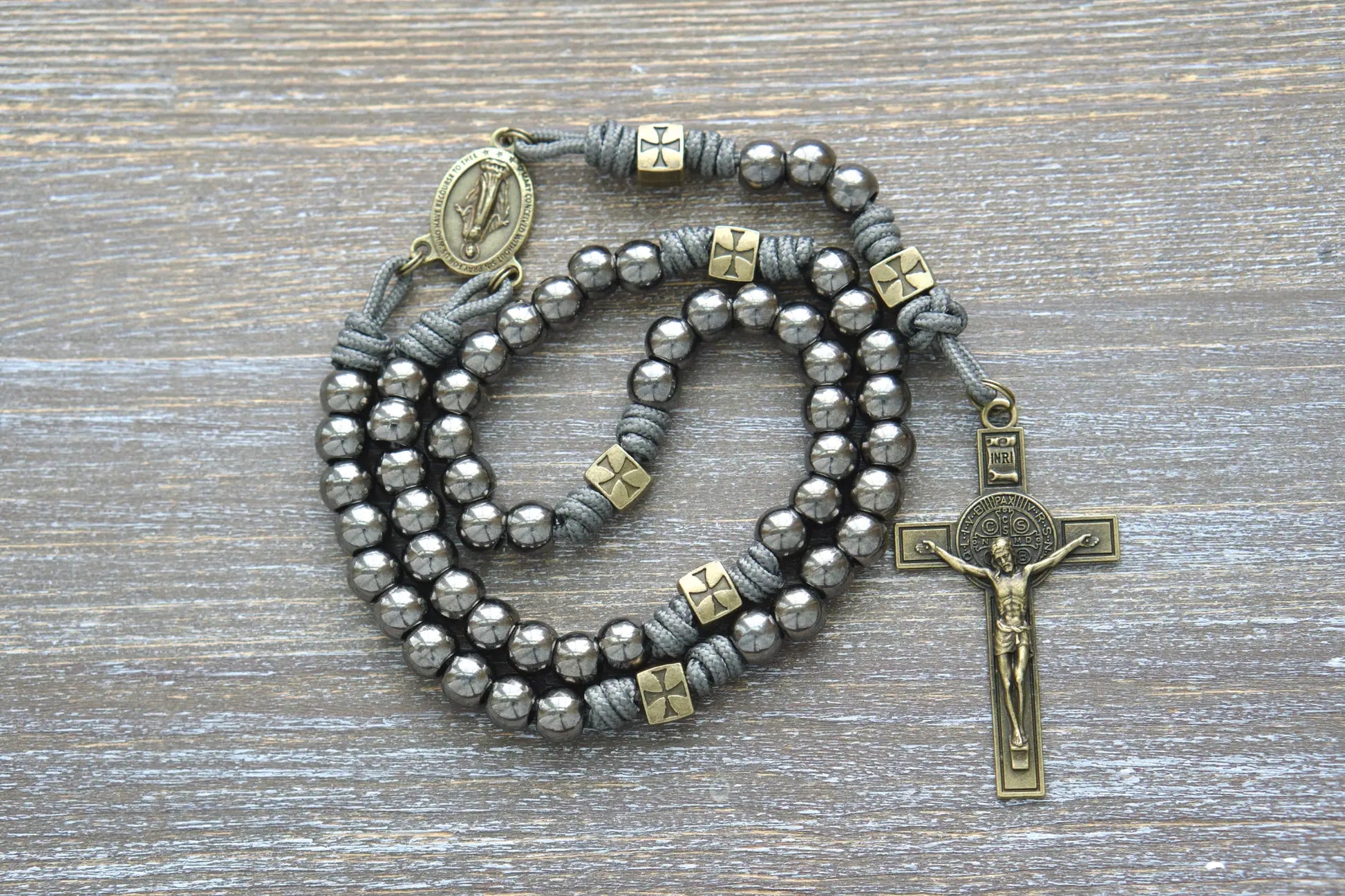A rugged metal paracord rosary featuring olive Our Father beads, gunmetal Hail Mary beads, a Miraculous Medal centerpiece, and a St. Benedict crucifix.