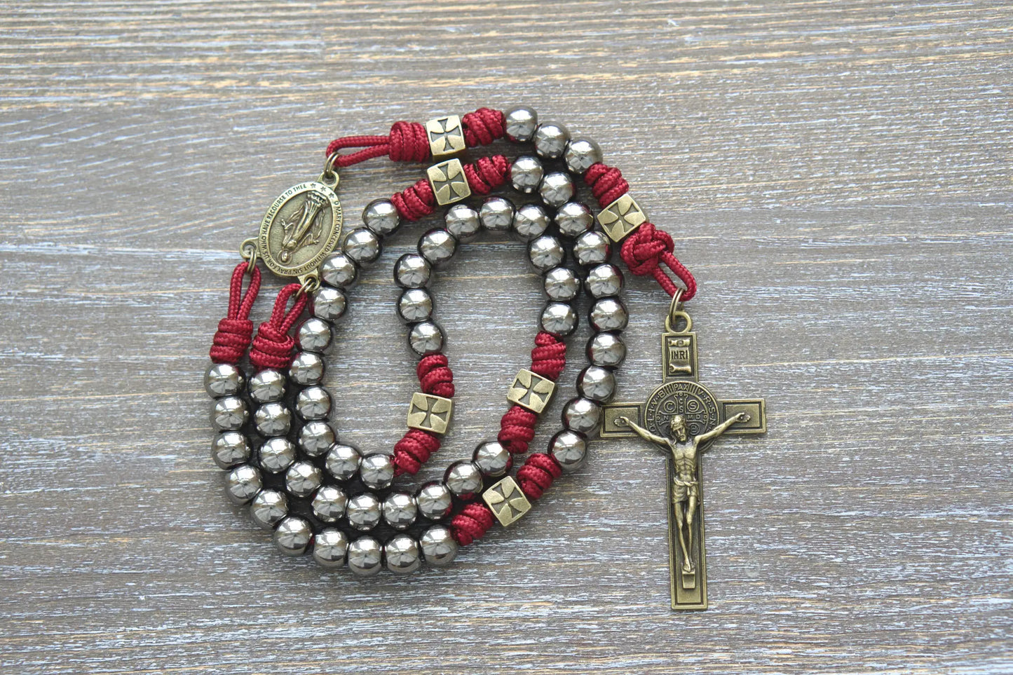 A handmade red paracord rosary featuring olive Our Father beads, gunmetal Hail Mary beads, a Miraculous Medal centerpiece, and a St. Benedict crucifix.