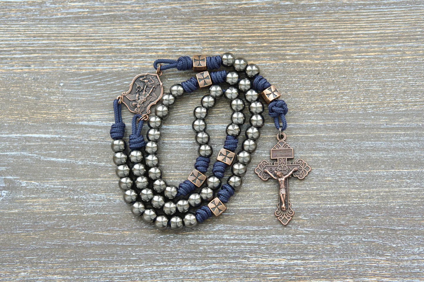 A navy blue paracord rosary featuring gunmetal metal Hail Mary beads, copper accents including a St. Michael centerpiece and copper Pardon crucifix.