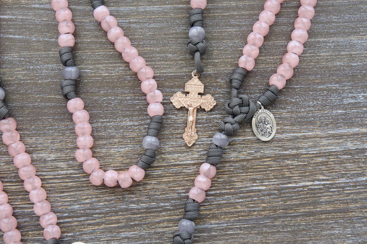 Ite Ad Joseph Paracord Rosary: A handmade, rosary featuring grey and pink colors, a rose gold Pardon crucifix, and a St. Joseph devotional medal.