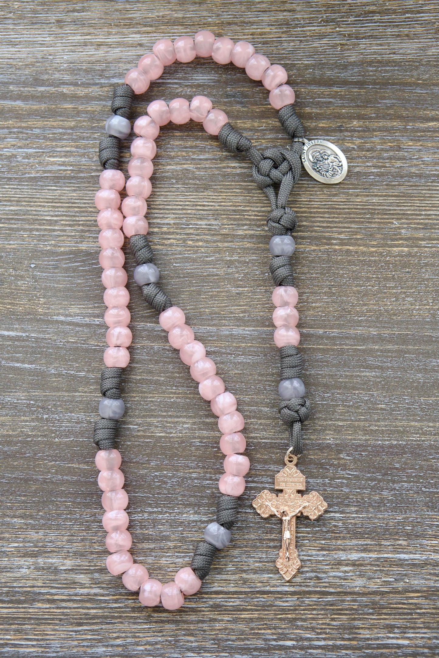 A woman's handcrafted 5-decade paracord rosary featuring grey and milky pink acrylic beads, a rose gold Pardon crucifix, and a St. Joseph devotional medal. Inspired by the traditional Catholic devotion to St. Joseph.