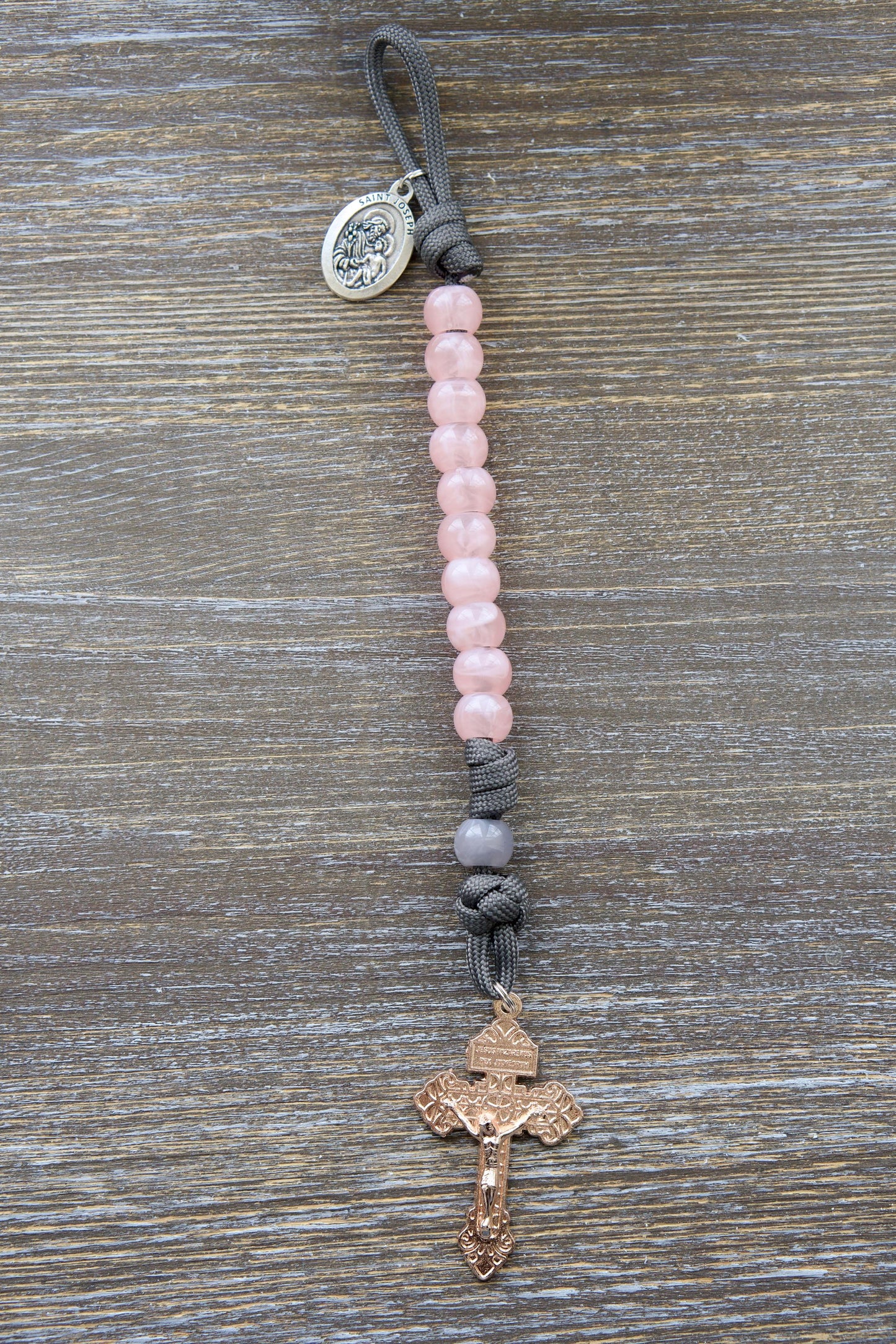 Single-decade grey paracord rosary featuring milky grey Our Father bead, contrasting pink Hail Mary beads, a rose gold Pardon Crucifix, and a St. Joseph devotional medal.