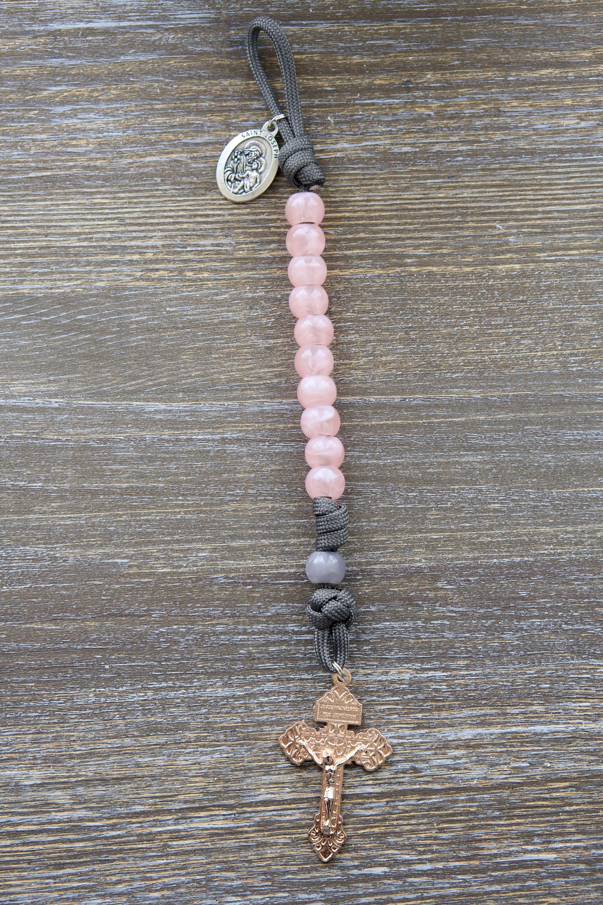 Single-decade grey paracord rosary featuring milky grey Our Father bead, contrasting pink Hail Mary beads, a rose gold Pardon Crucifix, and a St. Joseph devotional medal.