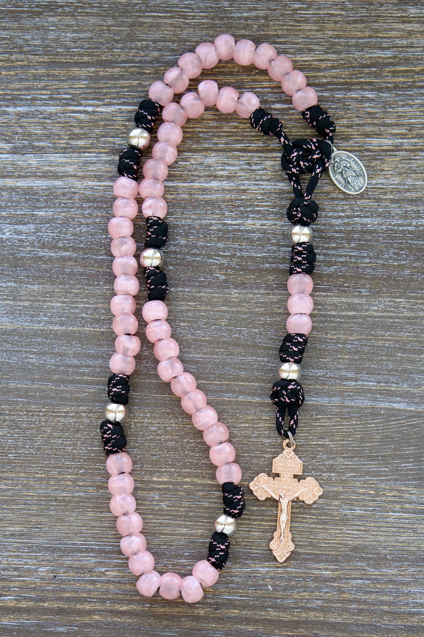 Our Lady Untier of Knots 5 Decade Paracord Rosary: A striking rosary featuring pink acrylic gemstone Hail Mary beads, silver Our Father beads, black and pink paracord, and an exclusive rose gold pardon crucifix. Includes the Our Lady Untier of Knots devotional medal.
