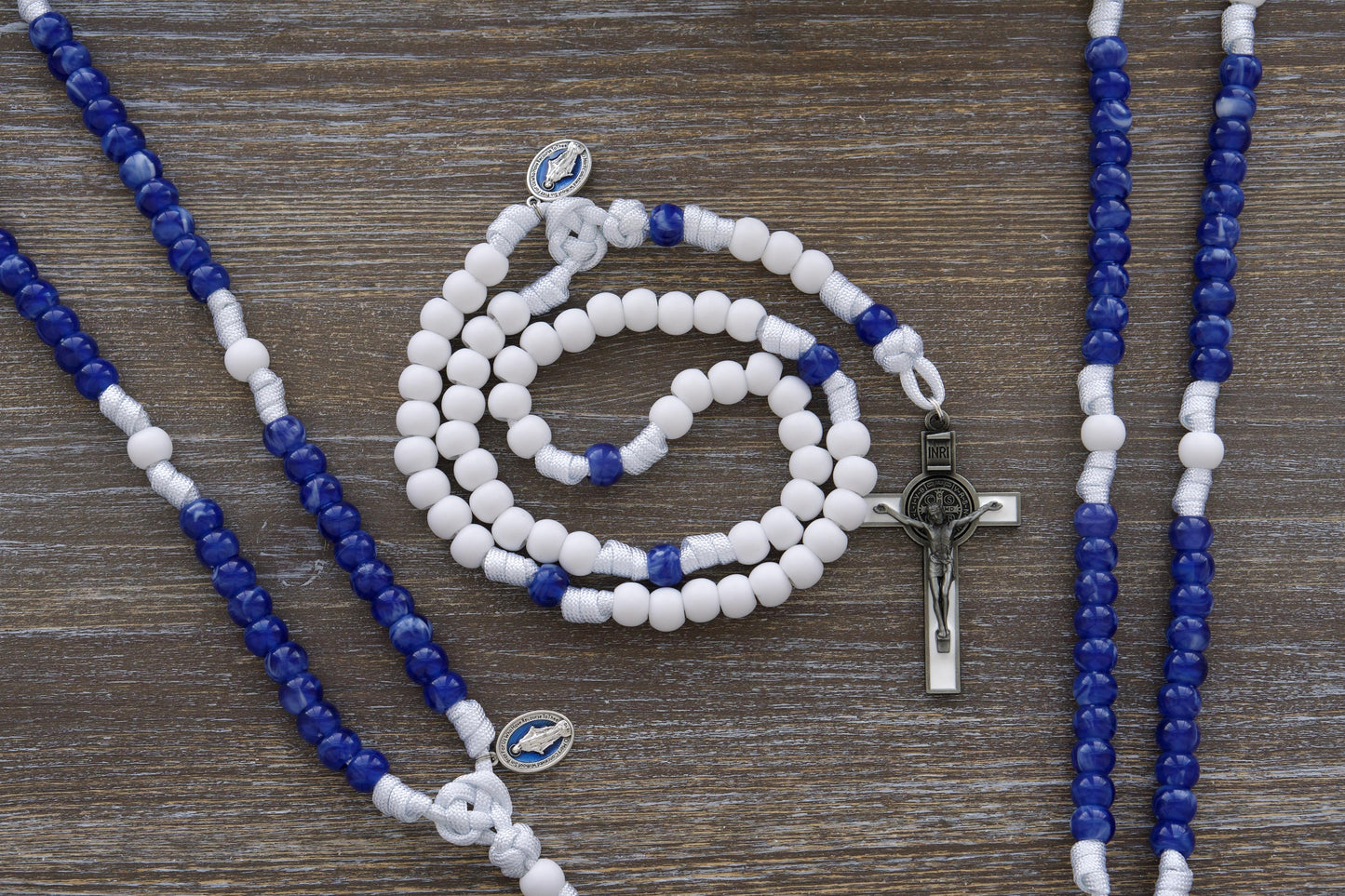 Our Lady's Mantle 5 Decade Paracord Rosary: A handcrafted rosary featuring white paracord, a blue enamel Miraculous Medal, and a white enamel St. Benedict crucifix. Two distinct style options offer 12mm matte white and miraculous blue acrylic beads, embodying devotion to Our Lady and spiritual protection.