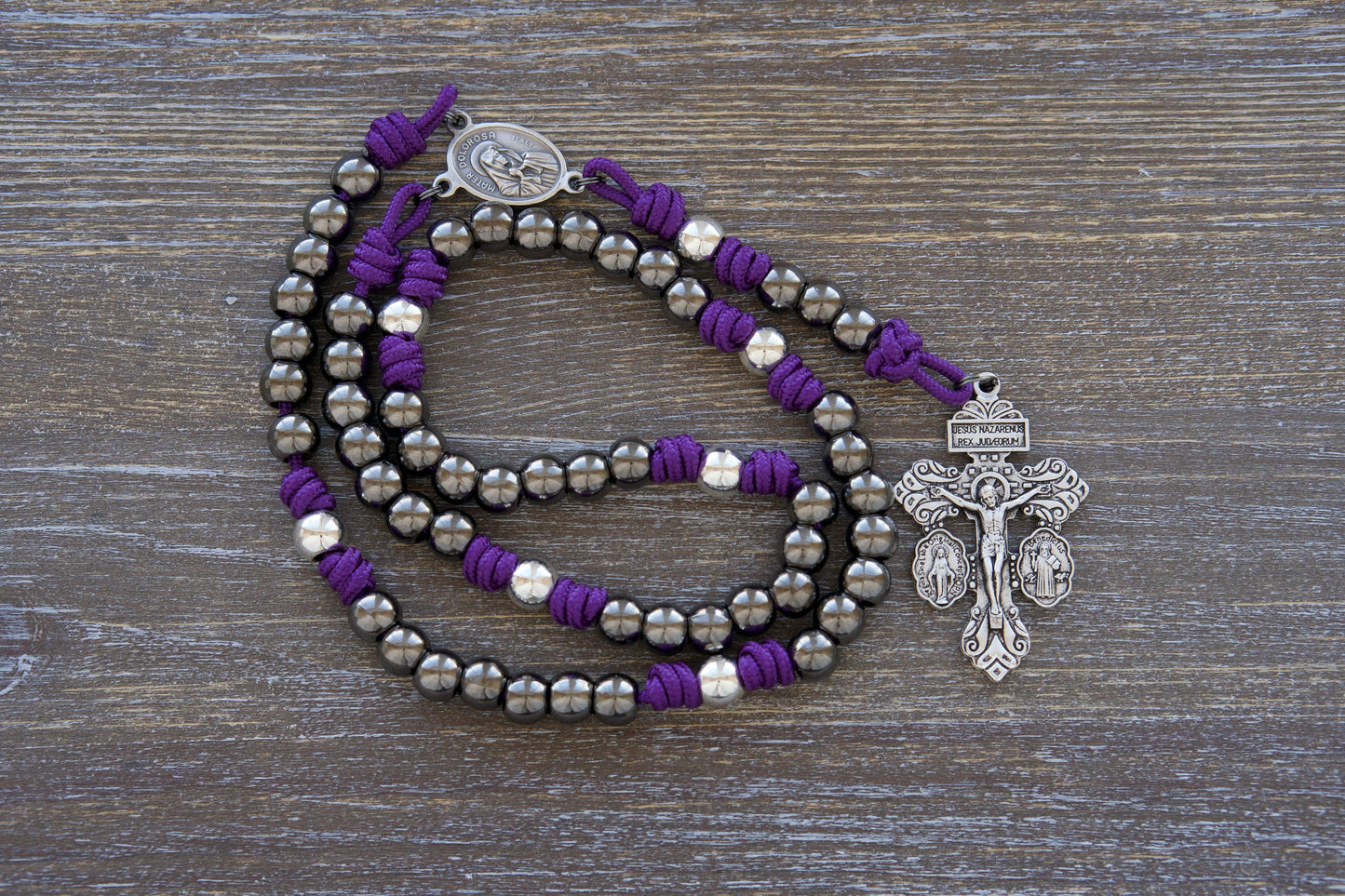A handcrafted '7 Sorrows Servite Rosary - Mater Dolorosa' chaplet in deep purple paracord. Features gleaming silver Our Father beads, gunmetal Hail Mary beads (10mm), a double-sided centerpiece medal depicting Mater Dolorosa and Ecce Homo, and a robust silver triple crucifix.