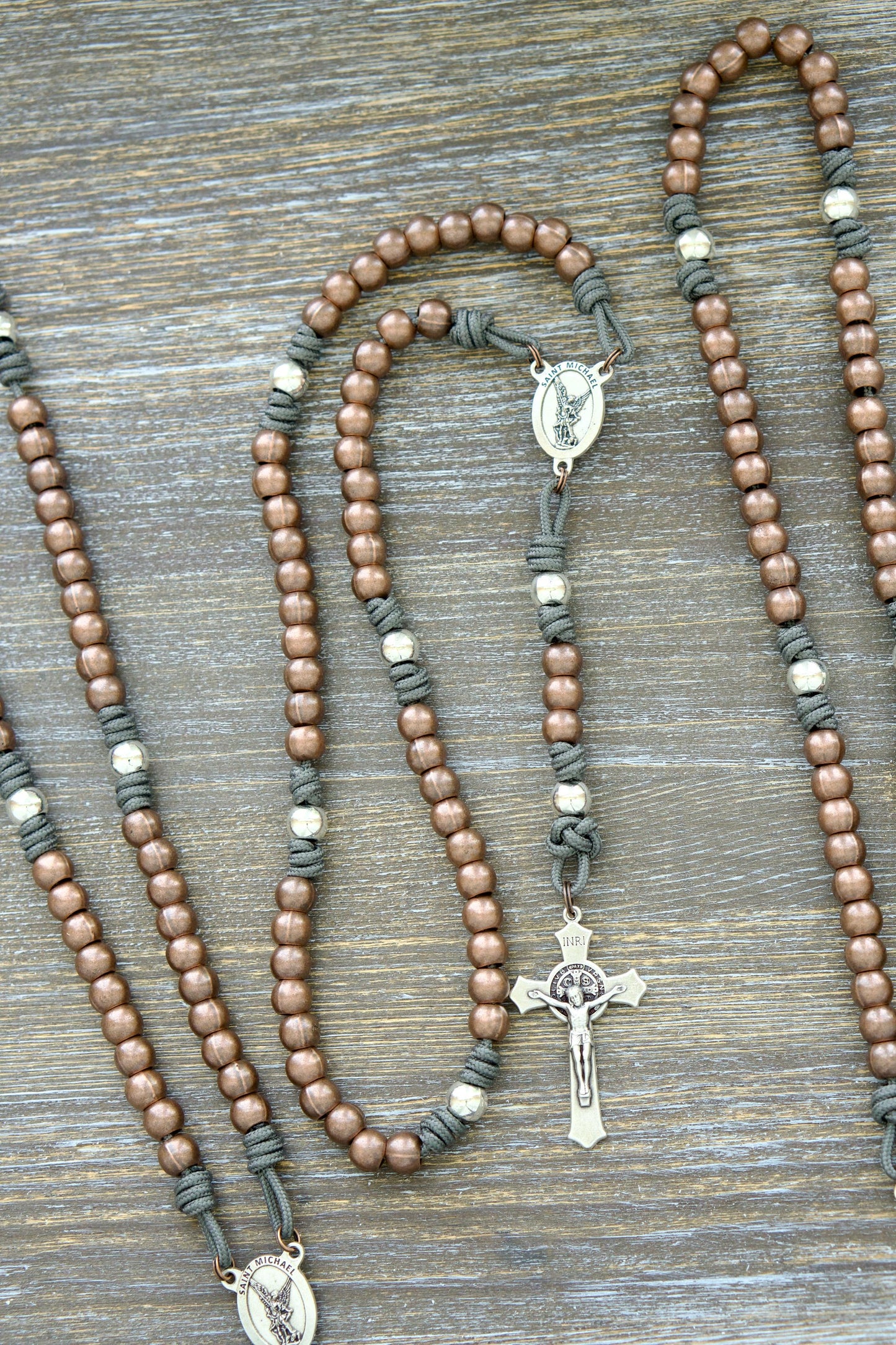 A handcrafted rosary featuring durable paracord (grey, black, green, or navy), silver Our Father beads, copper Hail Mary beads, a radiant St. Michael centerpiece medal, and a 2-inch St. Benedict crucifix.