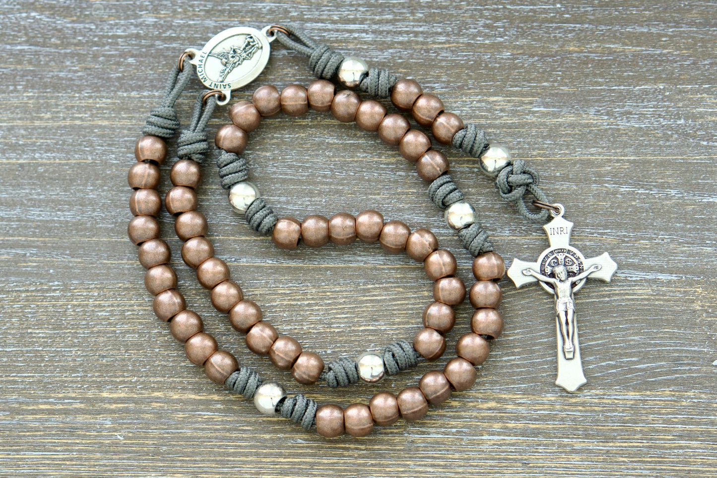 A handcrafted premium paracord rosary featuring durable grey, black, green, or navy blue paracord, 10mm alloy metal Hail Mary (copper) and Our Father (silver) beads, a radiant St. Michael centerpiece medal, and a 2-inch St. Benedict crucifix.