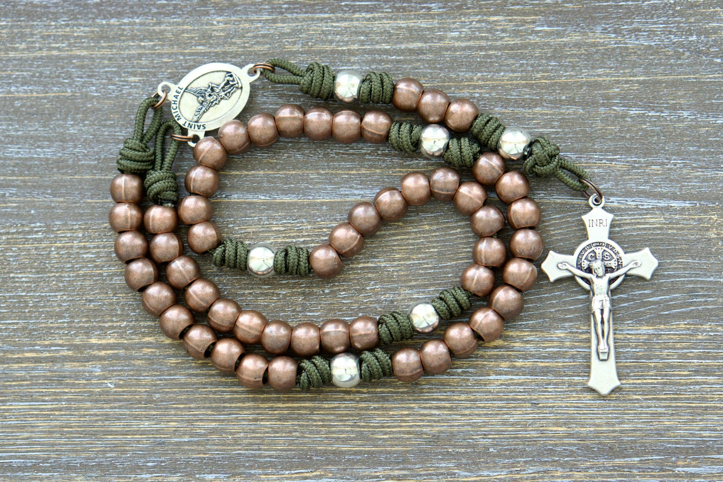 A rugged, handmade rosary featuring durable paracord in grey, black, green, or navy blue, accented with copper Hail Mary beads and silver Our Father beads. Centered around a St. Michael medal.