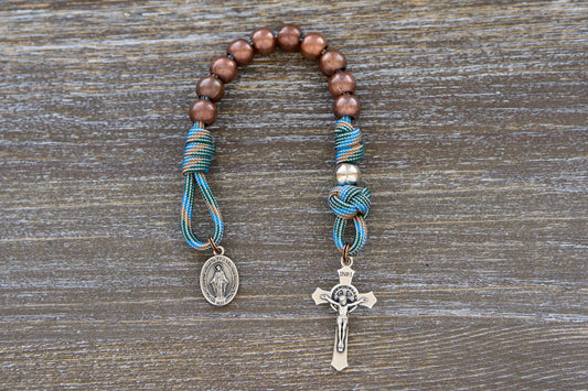 A handcrafted, pocket-sized paracord rosary designed for young boys. Features light blue and brown paracord, antique copper Hail Mary beads, silver Our Father beads, and a miniature Miraculous Medal crucifix.