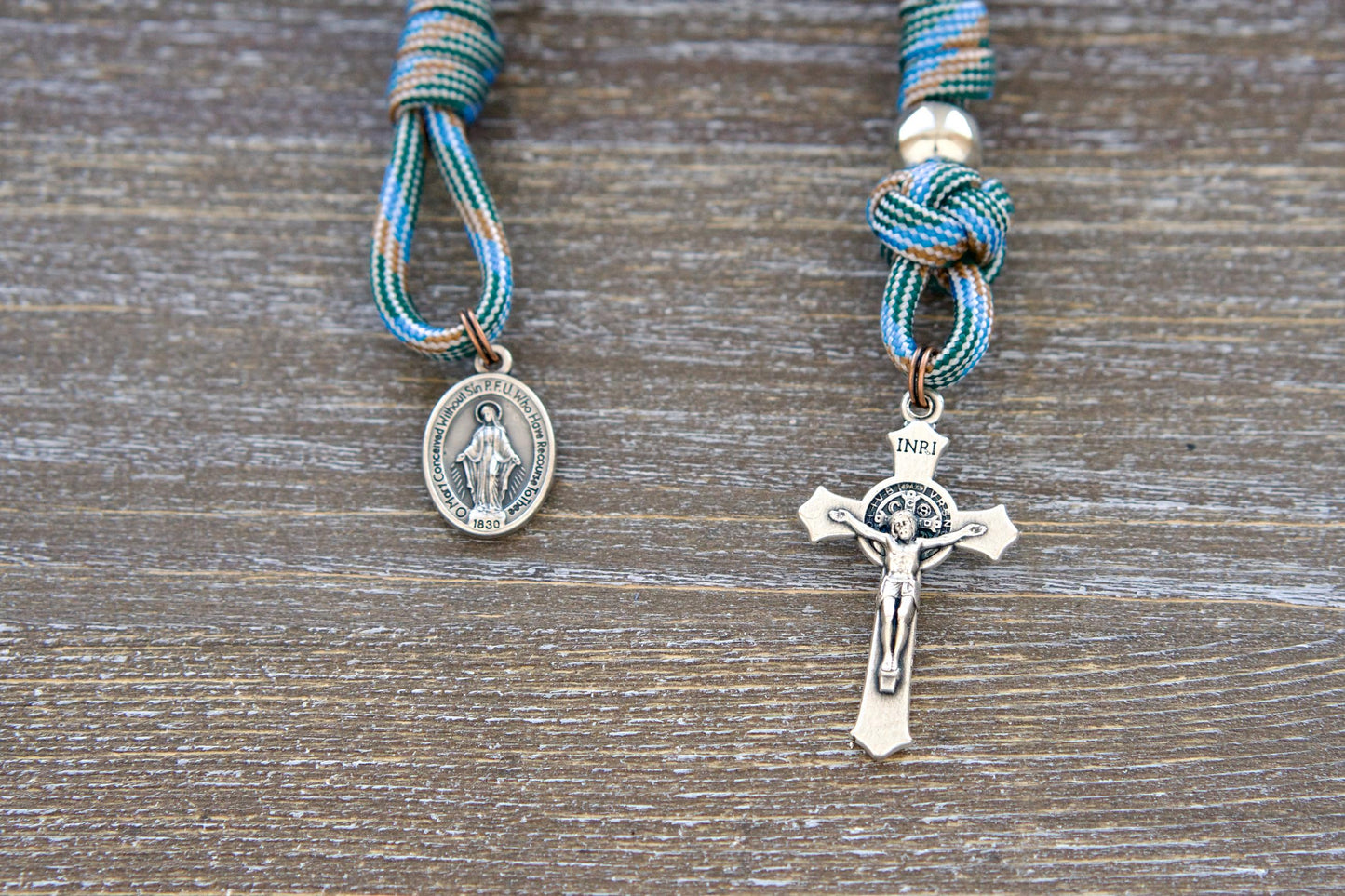 A close-up shot of the Unbreakable Faith Pocket Rosary, featuring light blue and brown paracord, antique copper Hail Mary beads, shiny silver Our Father beads, and a small Miraculous Medal pendant.