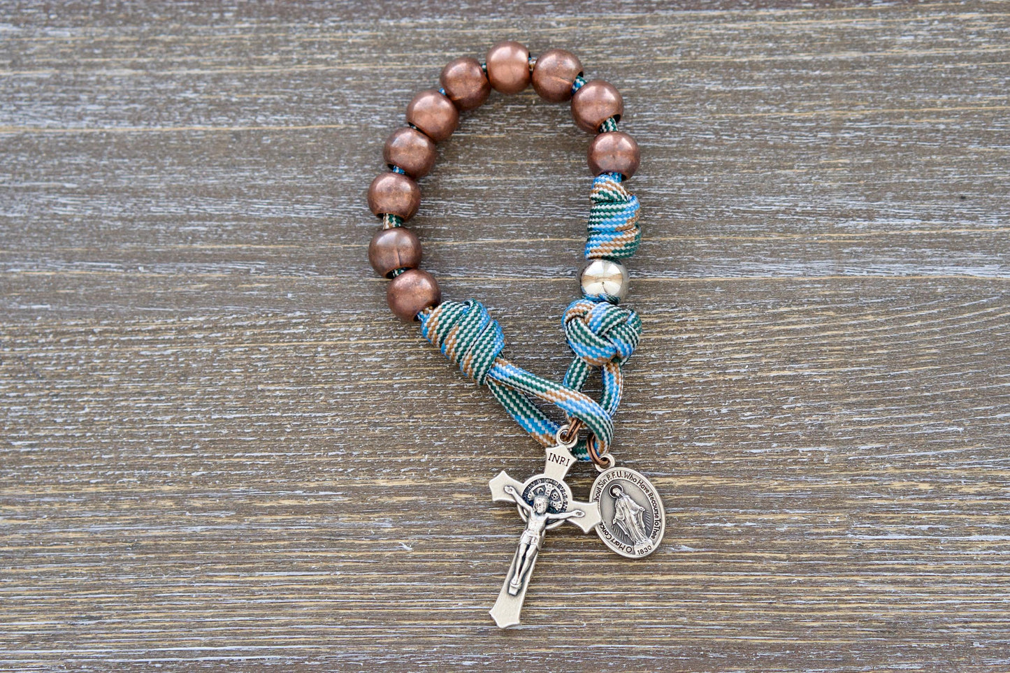 Handcrafted pocket paracord rosary in light blue and brown, featuring 10mm antique copper Hail Mary beads, silver Our Father beads, and a smaller Miraculous Medal crucifix for kids.