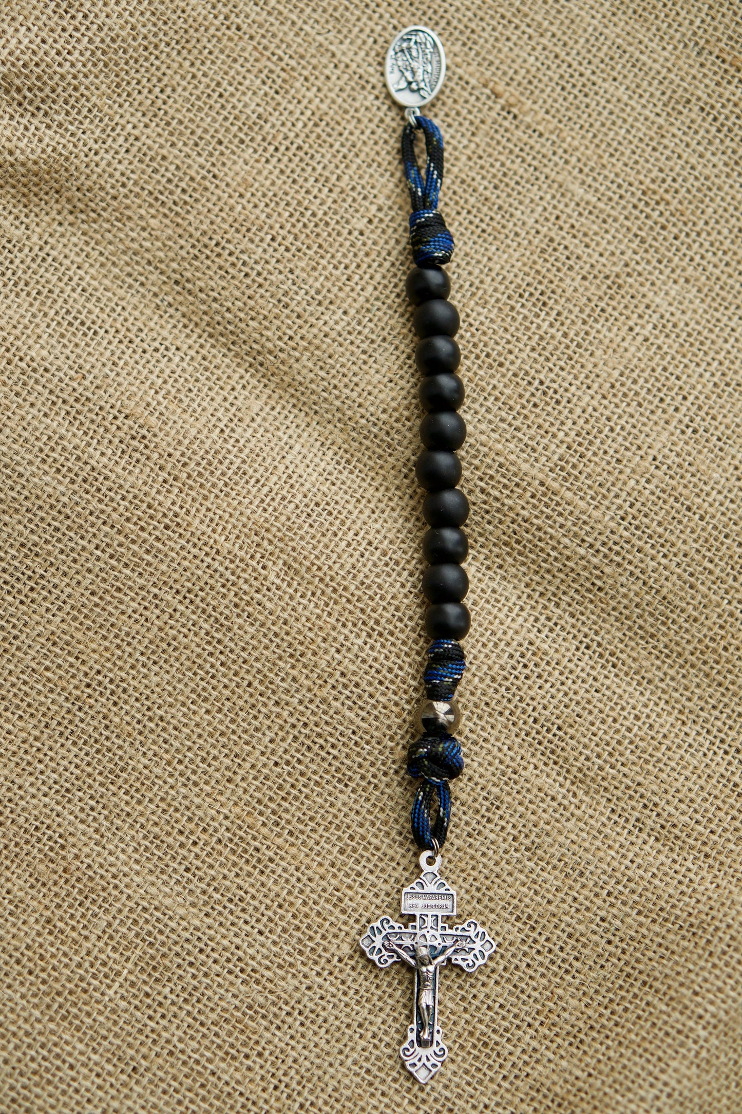 Thin Blue Line - 1 Decade Paracord Rosary featuring a blue/black camo design with black Hail Mary beads. This spiritual weapon is perfect for LEOs, complete with a gunmetal access bead and St. Michael devotional medal. A durable and unbreakable paracord rosary for Catholic gifts.