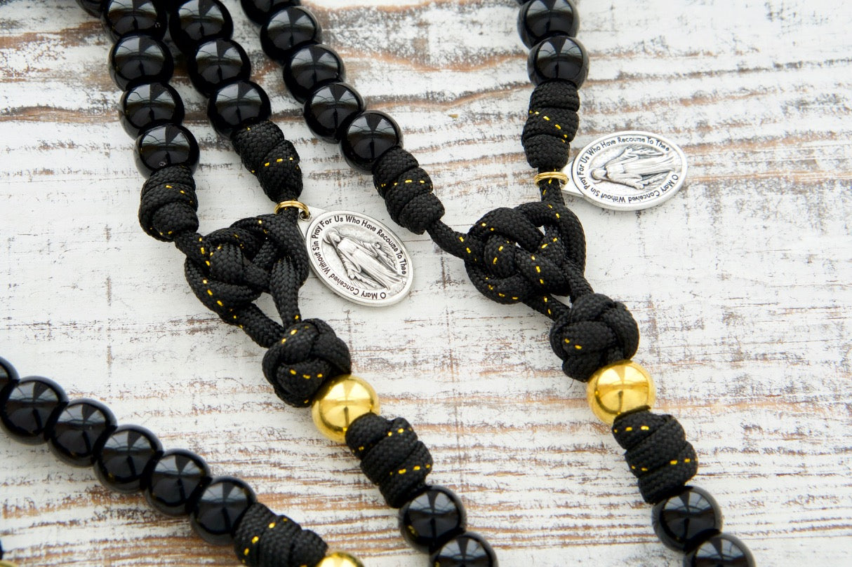 The Holy Servant - Black and Gold - 5 Decade Paracord Rosary: Elegant and devotional, featuring black paracord with gold ribbon, shiny black Hail Mary beads, golden Our Father beads, a large black enamel crucifix, and a miraculous medal. Perfect for personal prayer or as a thoughtful gift.