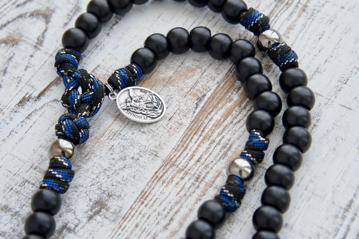Thin Blue Line - 5 Decade Paracord Rosary: A stylish, durable paracord rosary with a unique thin blue line camo rope design, perfect for law enforcement officers or those who appreciate the symbolism of the thin blue line. Featuring 12mm matte black Hail Mary beads, gunmetal Our Father beads, Pardon Crucifix, and devotional medal with St. Michael and Guardian Angel for spiritual guidance and protection.