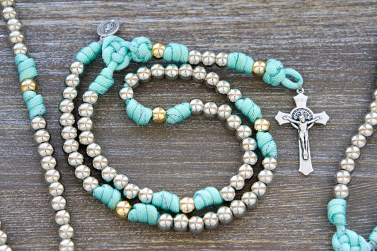 Teal blue paracord rosary with rainbow ribbon accents, silver and gold 10mm acrylic beads, a 2-inch St. Benedict crucifix, and a smaller Miraculous Medal charm. Designed as a durable and stylish 5-decade rosary for young Catholic girls.