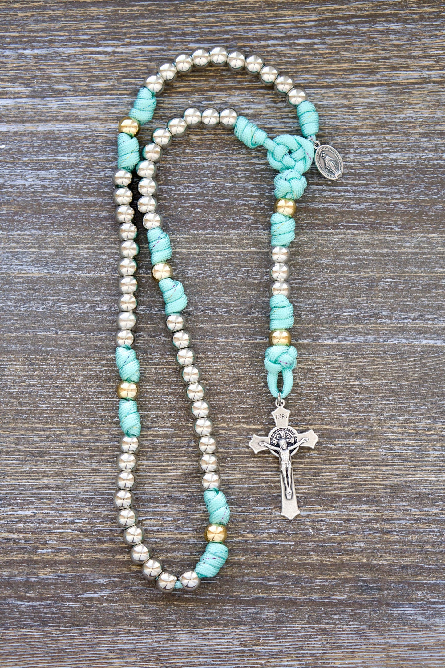 August Queen of the Heavens - Kid's 5 Decade Paracord Rosary: A teal blue paracord rosary with inlaid rainbow ribbon, royal silver and gold 10mm acrylic beads, a 2" St. Benedict crucifix, and a 3/4" Miraculous Medal.