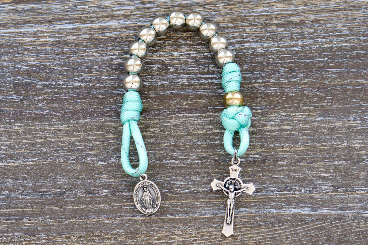 A single decade paracord rosary with 10mm acrylic beads, silver Hail Mary beads, a rose gold Our Father bead, a miniature St. Benedict crucifix, and a Miraculous Medal. Designed for children, it features durable paracord, perfect for backpacks or pockets