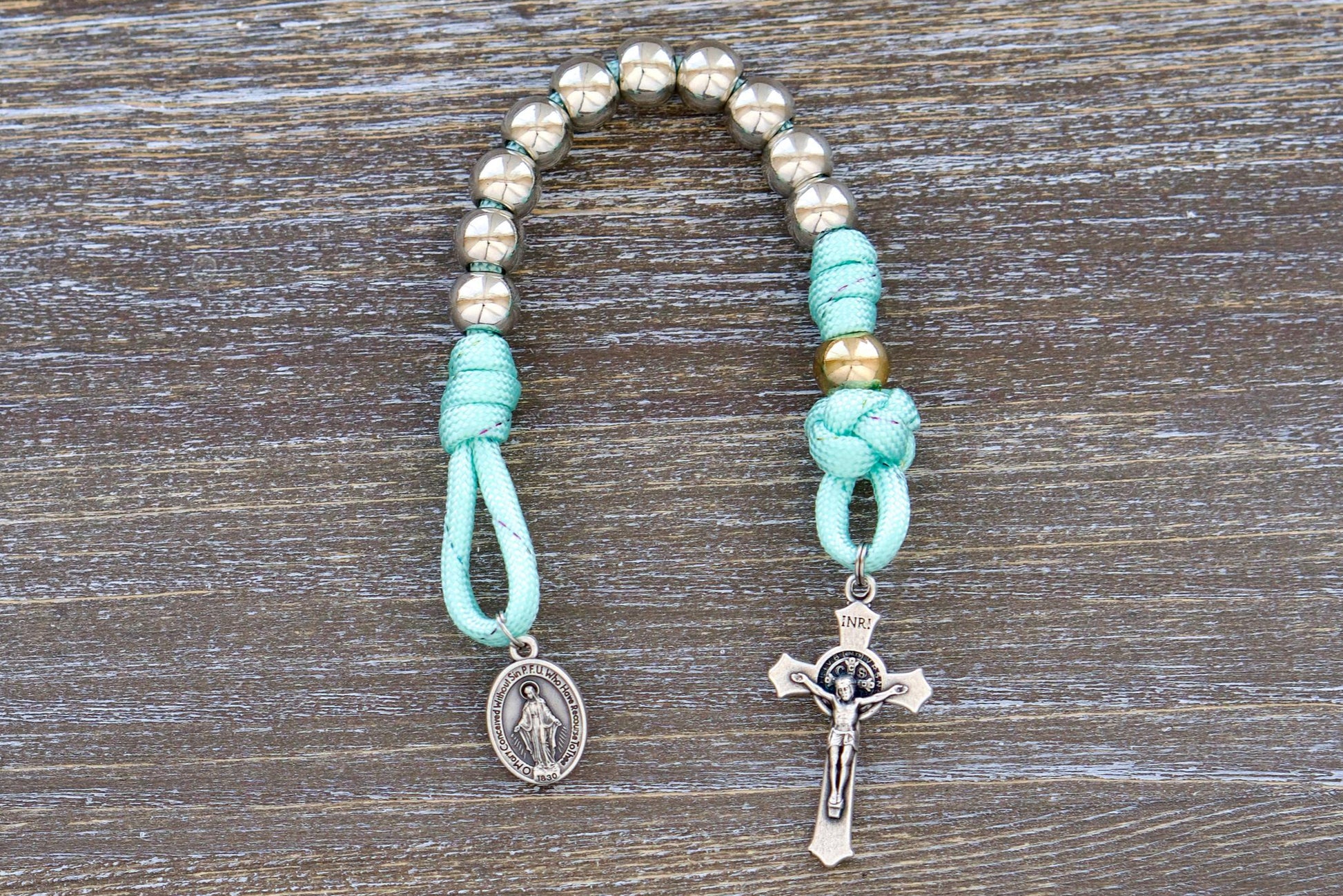 A single decade paracord rosary with 10mm acrylic beads, silver Hail Mary beads, a rose gold Our Father bead, a miniature St. Benedict crucifix, and a Miraculous Medal. Designed for children, it features durable paracord, perfect for backpacks or pockets