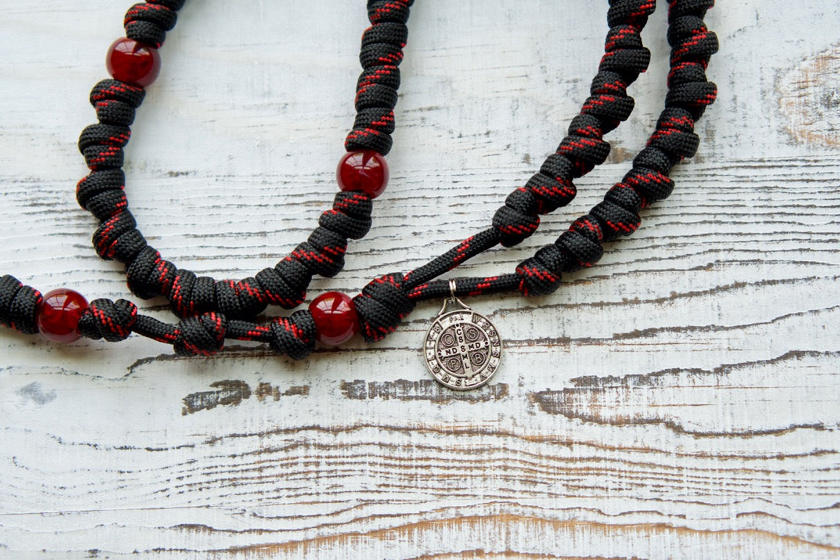 The Blood of Christ - Red and Black Knotted Rope Rosary, a durable and unbreakable paracord rosary handcrafted by our small Catholic family of 6. This powerful Catholic gift features bold red Our Father beads representing the Passion's blood, intricately knotted Hail Mary beads, a Pardon crucifix, and St. Benedict medal for spiritual significance.
