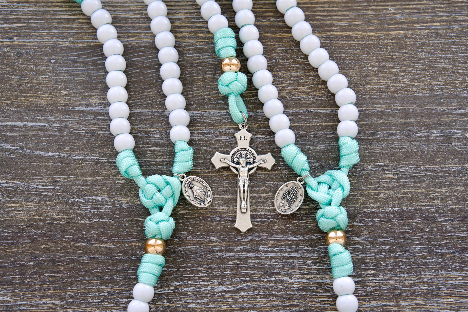 Close-up image of a mint green and white paracord rosary designed for children. Features durable 10mm acrylic beads, a rose gold Our Father bead accent, smaller Miraculous Medal, and a 2-inch St. Benedict crucifix.