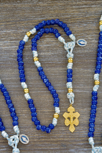 Divine Light 5 Decade Paracord Rosary: White and blue paracord, radiant gold Our Father beads, luminous 'Miraculous Blue' acrylic Hail Mary beads, blue enamel Miraculous Medal, and gold triple threat pardon crucifix. A handcrafted rosary for spiritual strength and prayer.