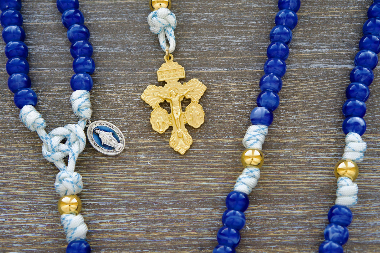 Divine Light 5-Decade Paracord Rosary: A handcrafted rosary featuring white paracord 550 with blue accents, radiant 12mm gold Our Father beads, exclusive luminous 'Miraculous Blue' acrylic Hail Mary beads, a blue enamel Miraculous Medal, and a gold Triple Threat Pardon crucifix.