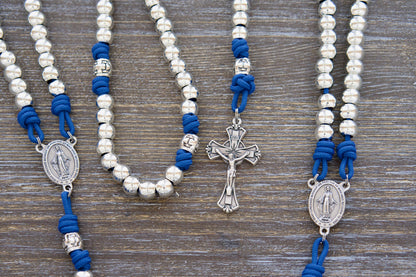 Divine Radiance Premium Metal Paracord Rosary: Close-up image showcasing lustrous 'miraculous blue' paracord, silver metal alloy Hail Mary beads, a shimmering silver Our Father cross barrel bead design, and the centerpiece Miraculous Medal and Crucifix. 