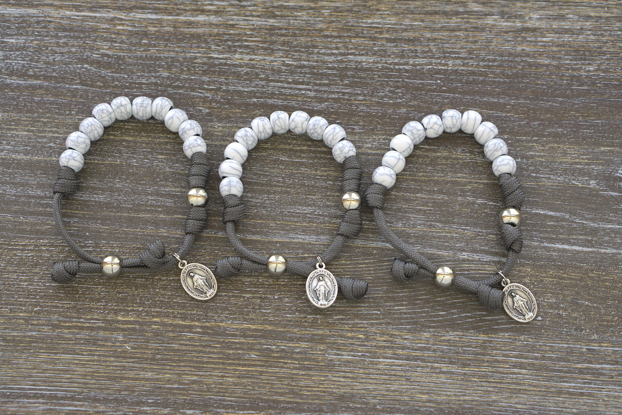 Faithful Fortress Kid's Rosary Bracelet, featuring a dark grey paracord rope, 'cracked ivory' 10mm beads for Hail Mary prayers, silver Our Father bead, and a Miraculous Medal.