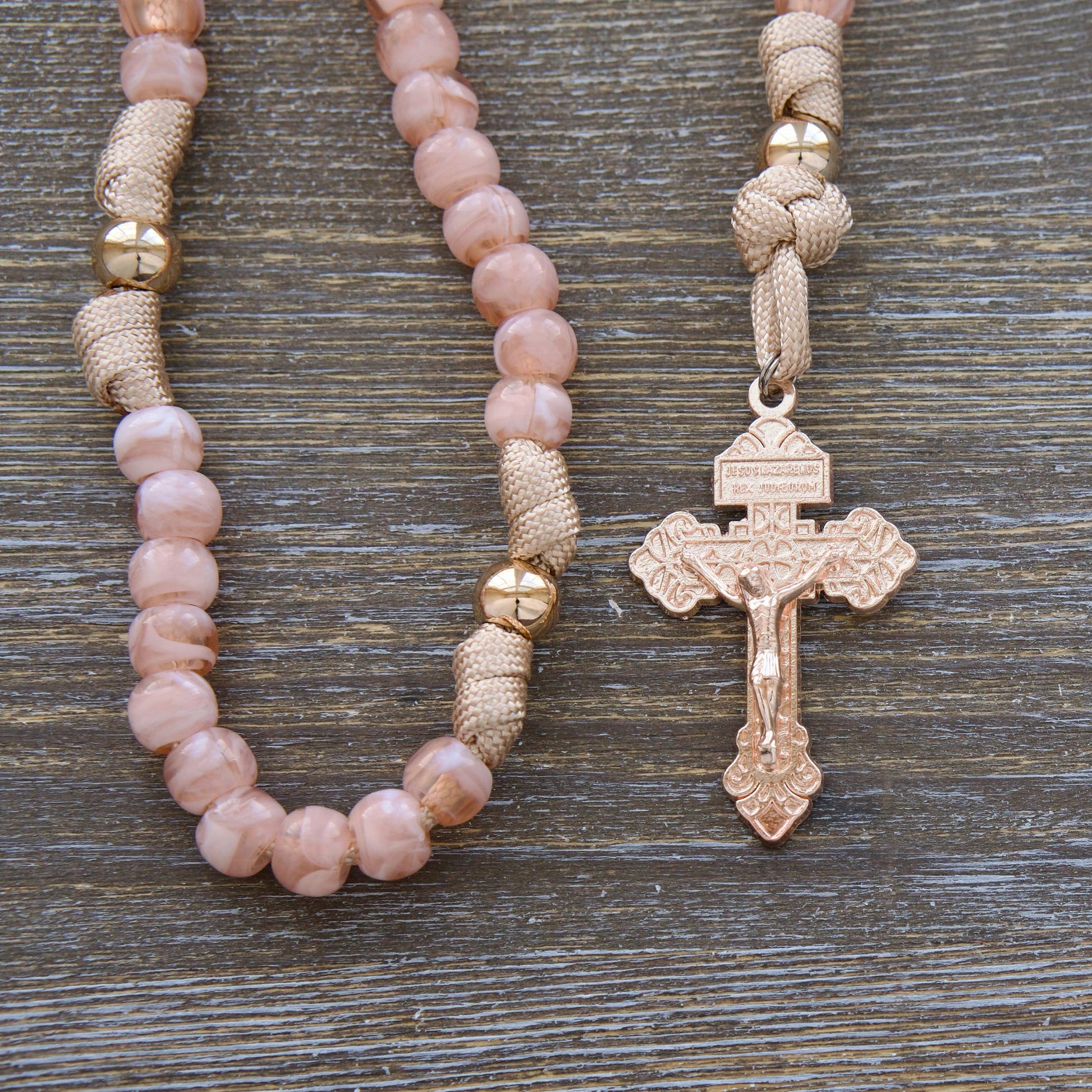 A child's paracord rosary named "Gate of Heaven", featuring rose gold/tan cord, milky pink Hail Mary beads, shiny gold Our Father beads, and a unique two-inch rose gold Pardon Crucifix. Includes a smaller Miraculous Medal devotional medal.