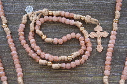 A child-sized paracord rosary, 'Gate of Heaven,' features rose gold and tan paracord interwoven with milky pink Hail Mary beads and shiny gold Our Father beads. It includes a unique two-inch rose gold Pardon crucifix and a smaller Miraculous Medal.