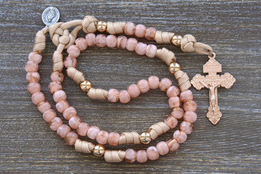 A child-sized paracord rosary named "Gate of Heaven," featuring rose-gold/tan paracord, milky pink Hail Mary beads, shiny gold Our Father beads, a custom two-inch rose gold Pardon Crucifix, and a smaller Miraculous Medal devotional medal.